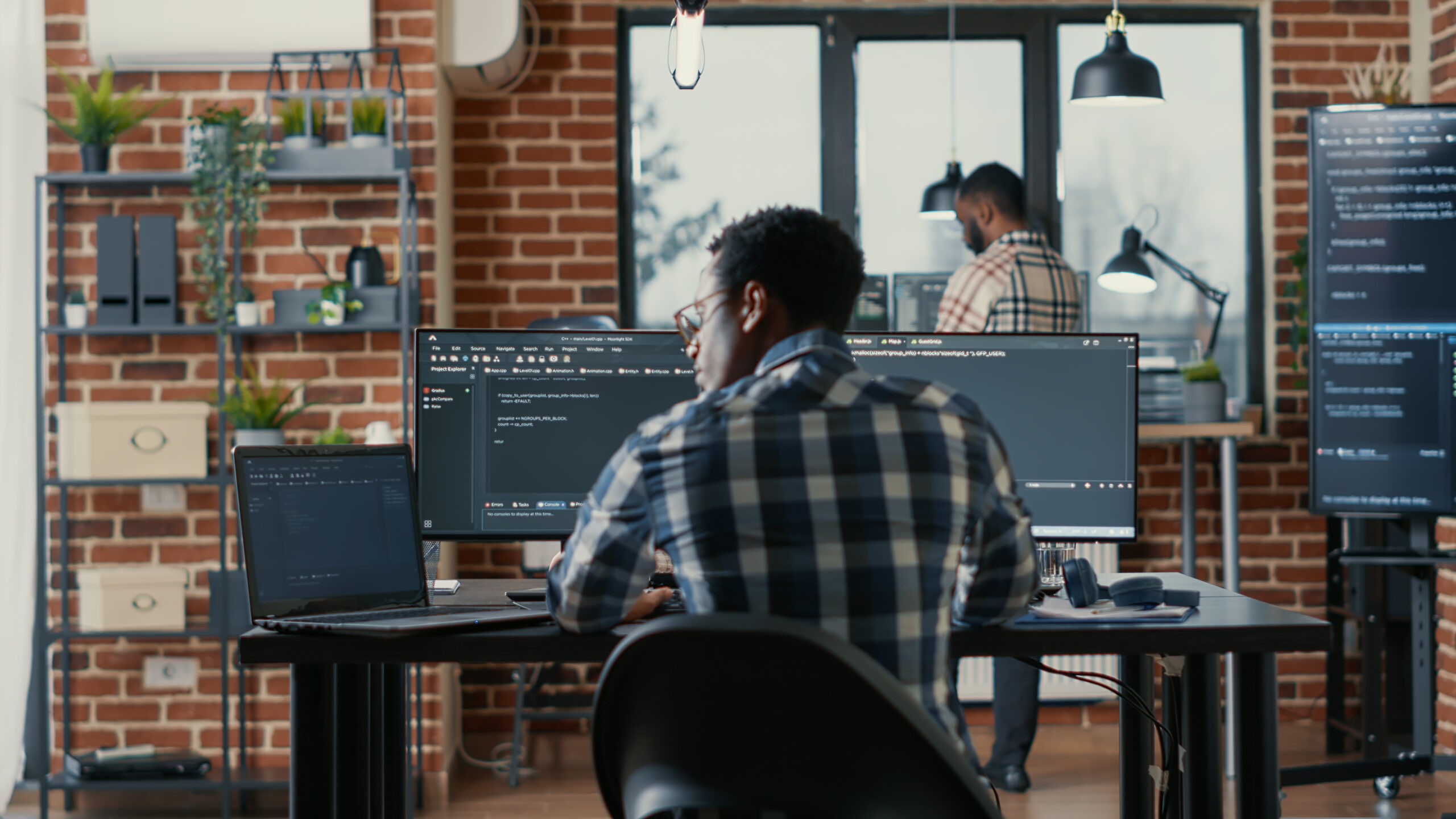 Programer coding on laptop sitting down at desk with computer screens parsing code